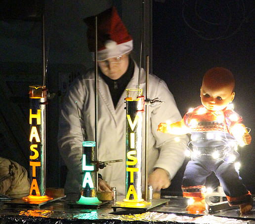 Chemiemitarbeiter mit Weihnachtsmütze steht am Experimentiertisch. Vor ihm stehen drei Raketen mit der Aufschrift: Hasta la vista.