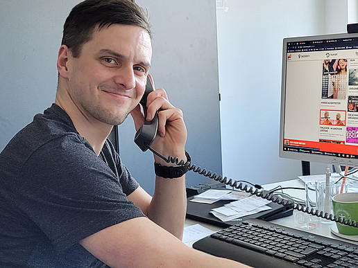 Rico Winkler lächelt in die Kamera, er sitzt am Schreibtisch, hält einen Telefonhörer in der Hand und auf seinem PC-Bildschirm im Hintergrund ist die Webseite von Radio Energy Sachsen eingeblendet.