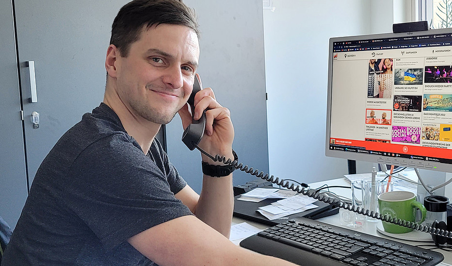 Rico Winkler lächelt in die Kamera, er sitzt am Schreibtisch, hält einen Telefonhörer in der Hand und auf seinem PC-Bildschirm im Hintergrund ist die Webseite von Radio Energy Sachsen eingeblendet.