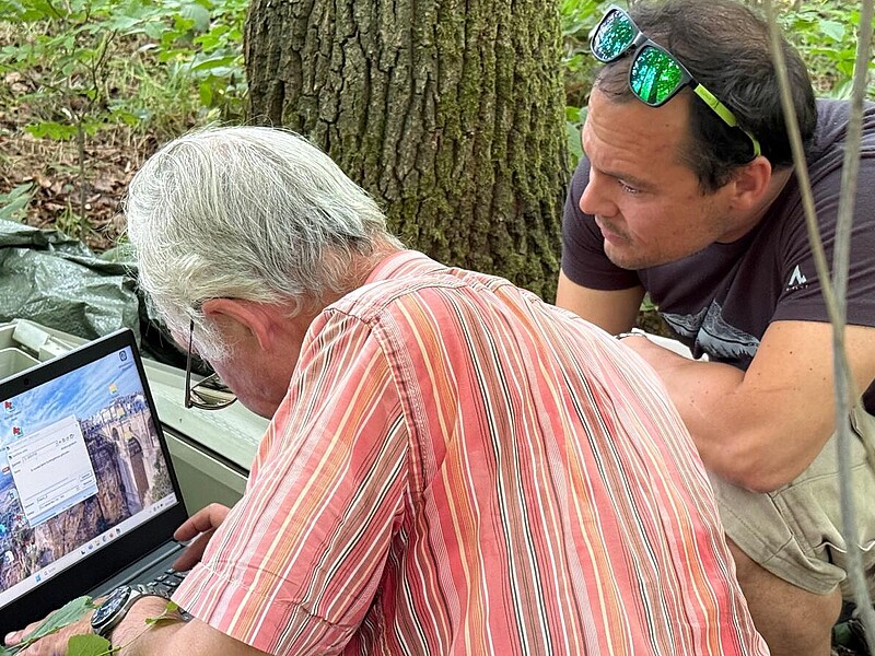 Prof. Hermann Heilmeier (TU Freiberg) und Prof. Oliver Wiche (HSZG) sitzen in einem Waldstück bei Freiberg am Boden vor einem geöffneten Metallkoffer. Heilmeier bedient einen Laptop, während Wiche aufmerksam über seine Schulter schaut. Im Hintergrund sind Bäume und Waldboden zu sehen. Die Szene zeigt das Auslesen eines Datenloggers im Rahmen einer europaweiten Messkampagne zum Bodenwasserhaushalt und Trockenstress in Bäumen, initiiert durch einen Aufruf der Gesellschaft für Ökologie im Mai 2025.