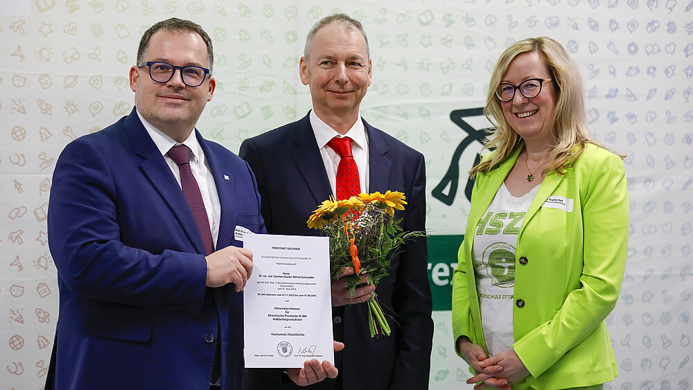 Der Rektor, Herr Schneider und Prorektorin Keil stehen vor einer HSZG-Pressewand. Der Rektor hält die Bestellungsurkunde zum Honorarprofessur in den Händen und Herr Schneider einen Blumenstrauß.