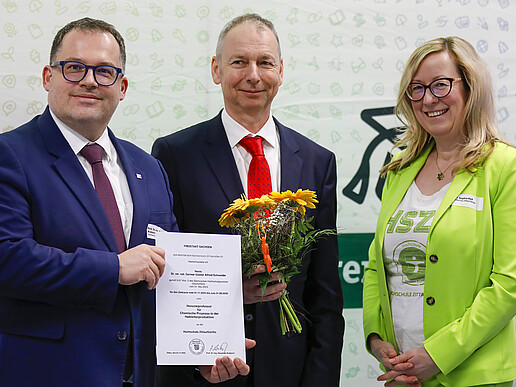 Der Rektor, Herr Schneider und Prorektorin Keil stehen vor einer HSZG-Pressewand. Der Rektor hält die Bestellungsurkunde zum Honorarprofessur in den Händen und Herr Schneider einen Blumenstrauß.