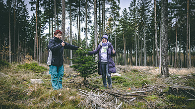Kanzlerin und Prorektorin im Wald beim Einpflanzen, beide zeigen ihren gehobenen Daumen.