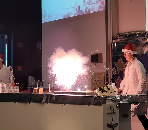 Zwei Laboranten mit Weihnachtsmützen experimentieren auf der Bühne. In der Mitte des Tisches leuchtet ein Feuerwerk.