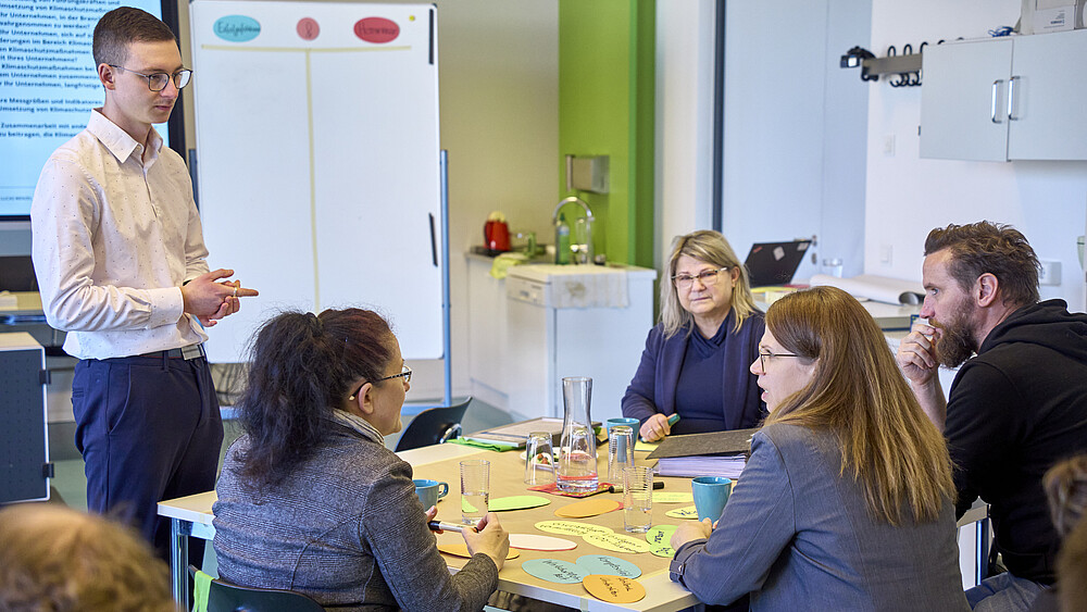 An einem Projekttisch sitzen Vertretende von Unternehmen. Ein HSZG-Ansprechpartner steht neben einem weißen Flipchart.