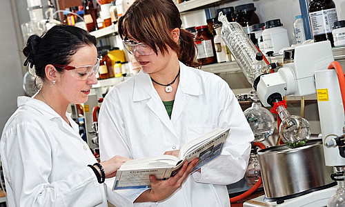 Laborantinnen in Schutzkleidung prüfen im Labor einen Text aus einem Lehrbuch und beraten darüber