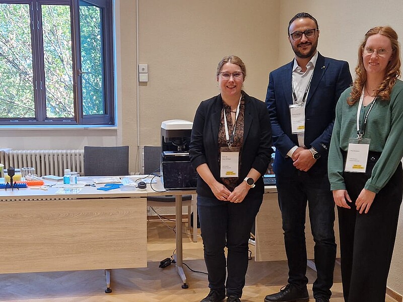 Prof. Dr. Elisa Wirthgen, Ali Al Hamad und Pia Schubert stehen vor dem HSZG-Stand mit Mikroskop und Informationsmaterialien zur biomedizinischen Forschung.