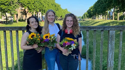Drei junge Frauen stehen auf einer Brücke, lächeln in die Kamera und halten Blumensträuße in den Händen, die sie für ihren Deutsch-Russischen-Doppelabschluss erhalten haben.. 